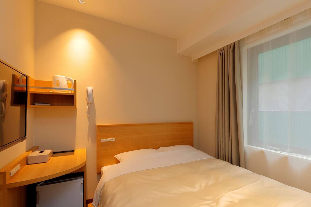 A hotel room with a bed, a desk with a telephone, a mirror, and a window with curtains.