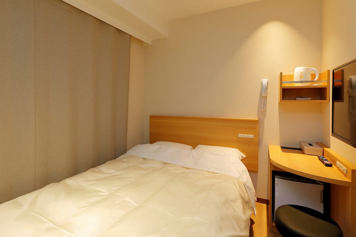 A hotel room with a bed, a desk with a chair, a telephone, and a small shelf with a cup and a remote control.