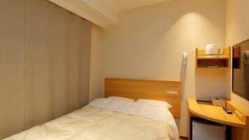 A hotel room with a bed, a desk with a chair, a telephone, and a small shelf with a cup and a remote control.