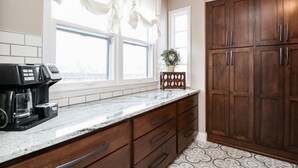 A kitchen with a coffee maker, wooden cabinets, a window with curtains, and a vase with flowers.
