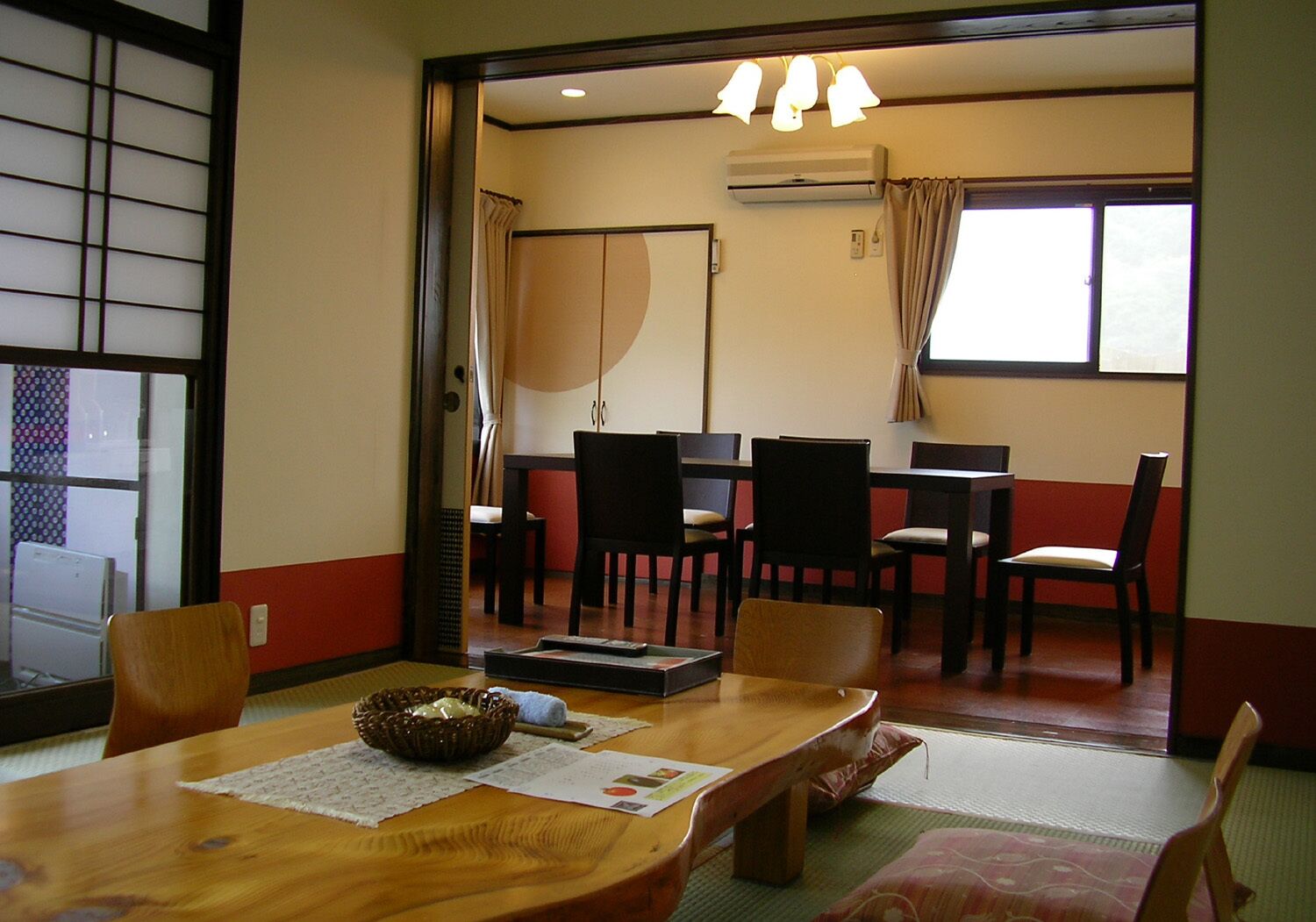 A traditional Japanese-style room with tatami flooring, a low table with a woven basket, and a sliding door leading to a dining area with a table and chairs.