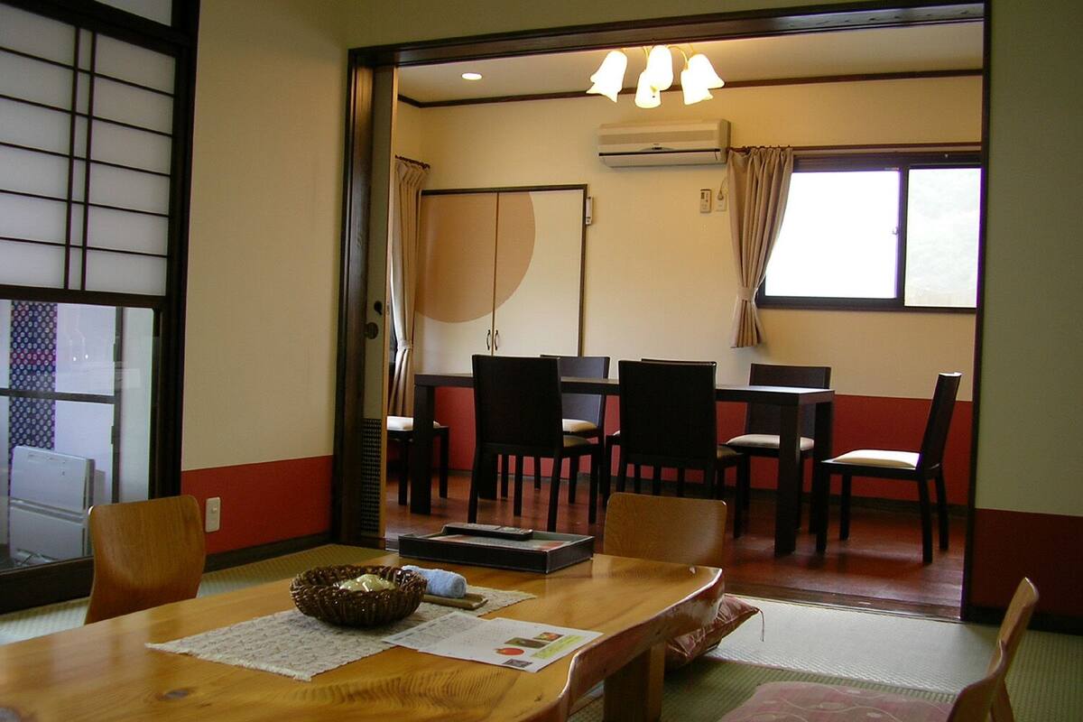 Um quarto tradicional em estilo japonês, com piso de tatame, uma mesa baixa com um cesto trançado e uma porta deslizante que dá para uma área de jantar com mesa e cadeiras.