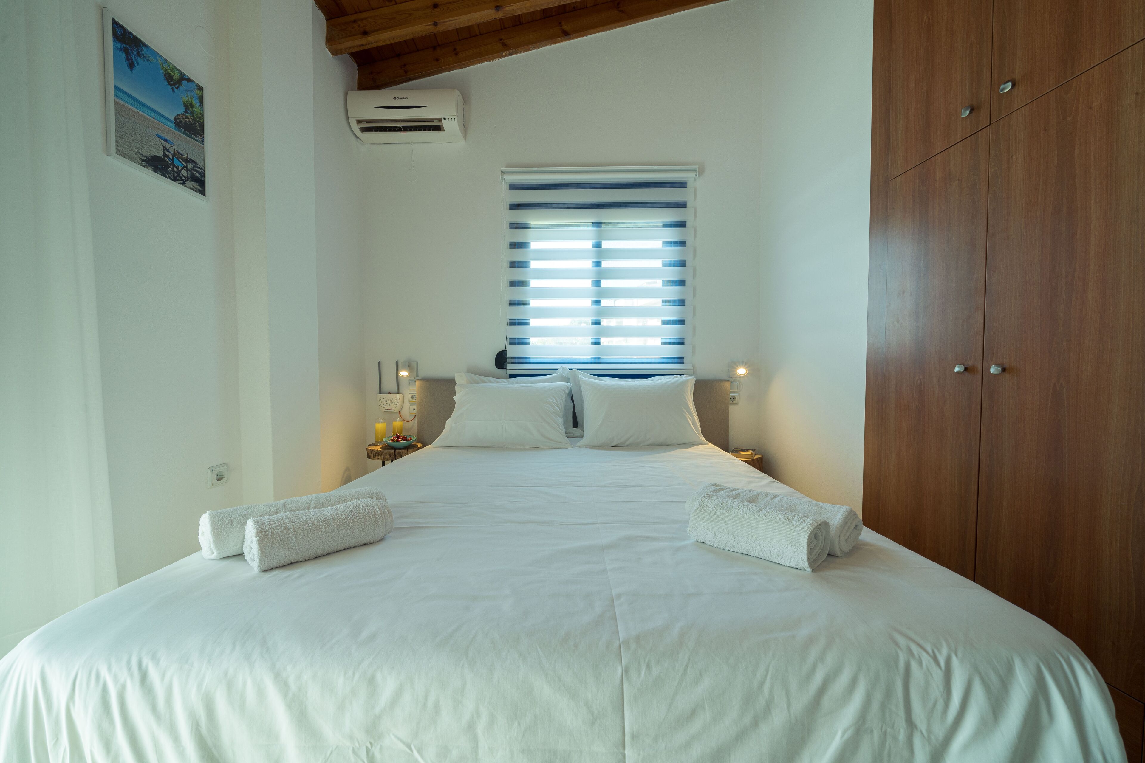 A bedroom with a large bed, white linens, a window with blinds, a wooden wardrobe, and an air conditioning unit.