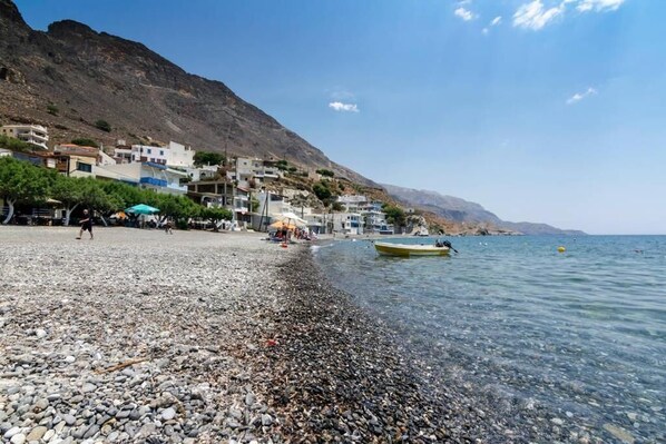 A pebble beach with buildings on a hillside, a boat on the water, and a clear sky. - Sunset home crete 3 (treis ekklisies)