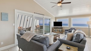 A spacious living room with a large gray sofa, a coffee table, and a television. There is a ceiling fan, a large window with a view, and a door leading to a balcony. - Omni Resort - Gorgeous Top Floor Penthouse with Oceanfront Views - Shipwatch! (Fernandina Beach)