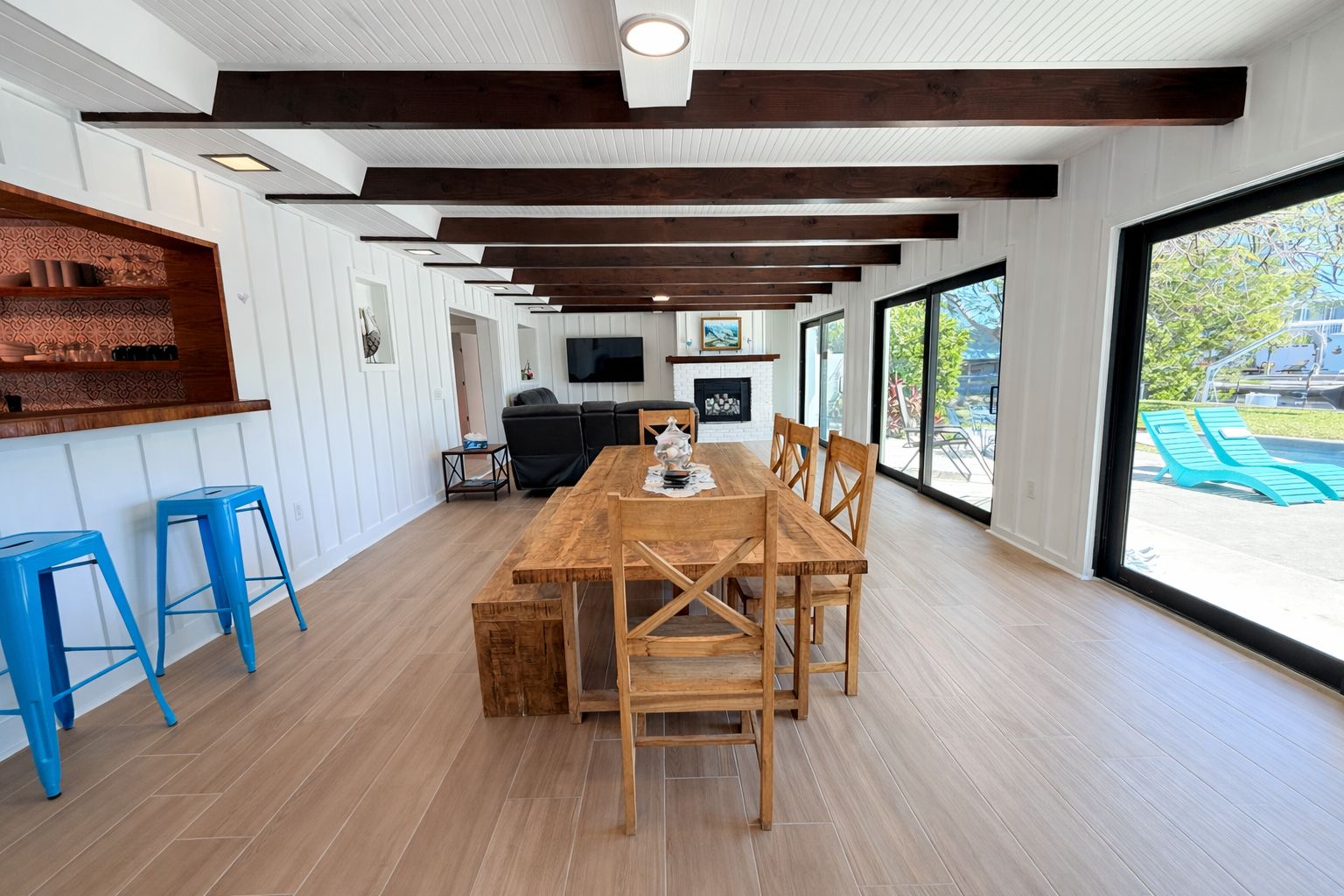 A spacious dining area with a wooden table and chairs, a TV mounted on the wall, and a view of the outdoors through large windows.