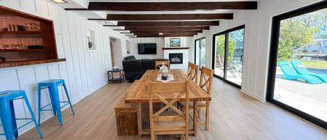 A spacious dining area with a wooden table and chairs, a TV mounted on the wall, and a view of the outdoors through large windows.
