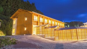 Un edificio grande de madera con múltiples ventanas y techo inclinado, rodeado por una valla y árboles.
