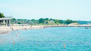 Una playa con gente nadando y tomando el sol, un muelle y casas al fondo.