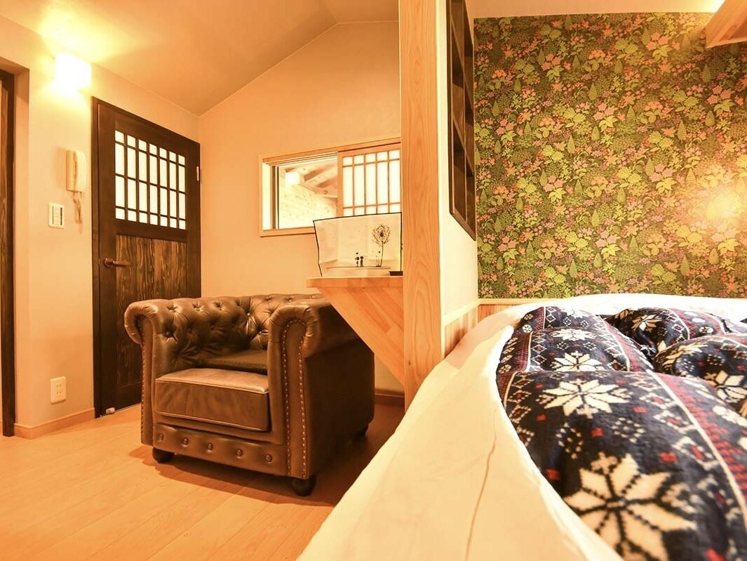 Ein Zimmer mit einem Bett, einem Ledersessel, einem Holztisch, einem Spiegel und einer Wand mit Blumenmuster.