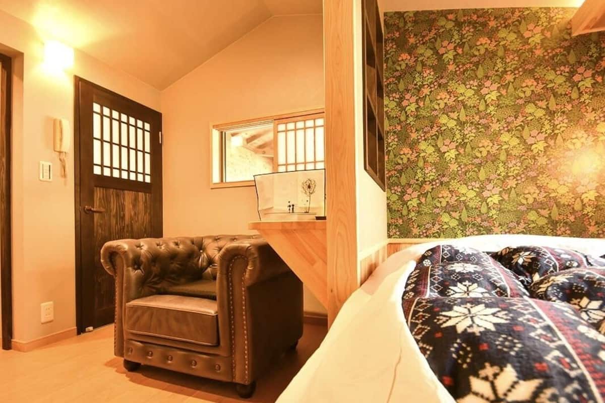 A room with a bed, a leather armchair, a wooden desk, a mirror, and a wall with a floral pattern.