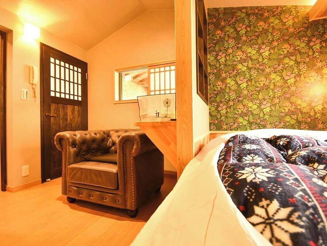 Una habitación con una cama, un sillón de cuero, un escritorio de madera, un espejo y un papel tapiz con motivos florales.