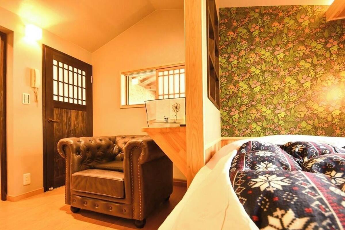 A room with a bed, a leather armchair, a wooden desk, a mirror, and a floral wallpaper.