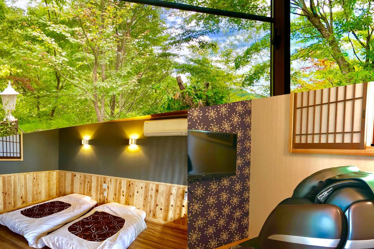 A room with a wooden floor, two beds with white and brown bedding, a wall-mounted TV, and a view of trees through a window.