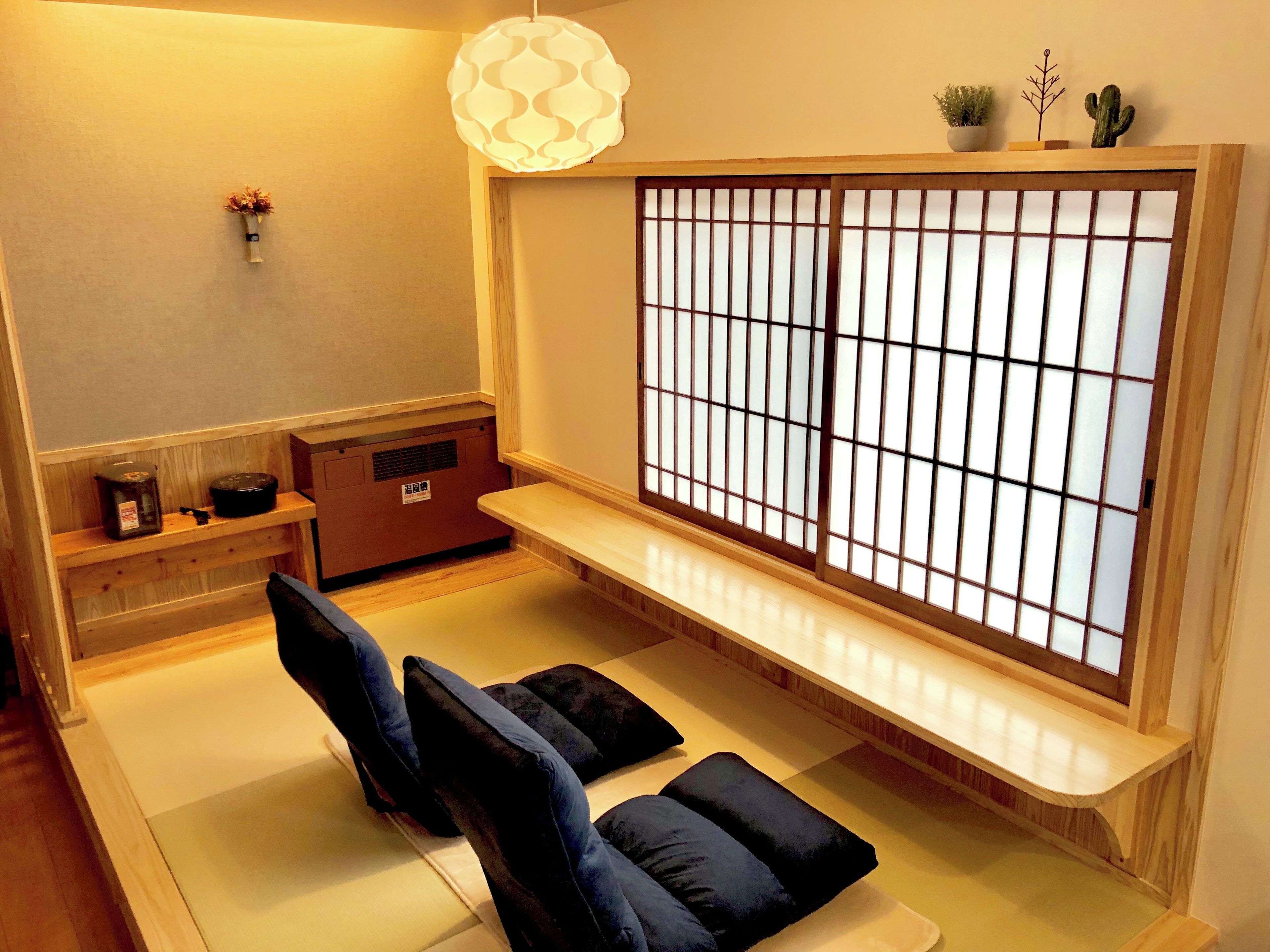 Una habitación japonesa tradicional con suelo de tatami, una mesa de madera baja, un banco acolchado, un celosía de papel shoji y una lámpara colgante.