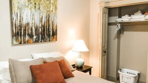 Een netjes opgemaakte hotelkamer met een bed, kussens, een lamp, een schilderij en een kledingkast met daarin hangende kleding.
