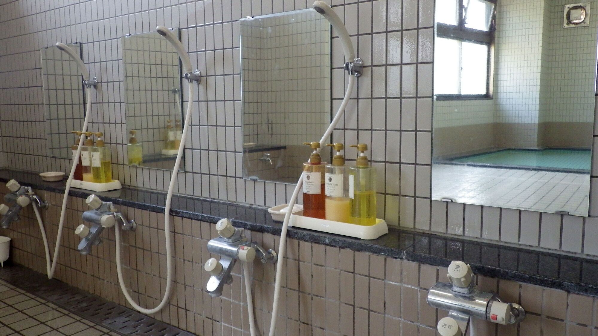 A tiled bathroom with three mirrors, multiple faucets, and toiletries.