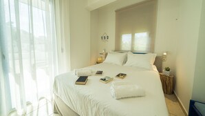 A neatly made bed with white linens, a bedside table with a lamp, and a window with sheer curtains. - Sunset home crete 2 (Τρεις Εκκλησίες)