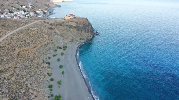 An aerial view of a coastal area with a small building on a cliff overlooking the sea, a road leading to a settlement, and a rocky shoreline. - Sunset home crete 2 (Τρεις Εκκλησίες)
