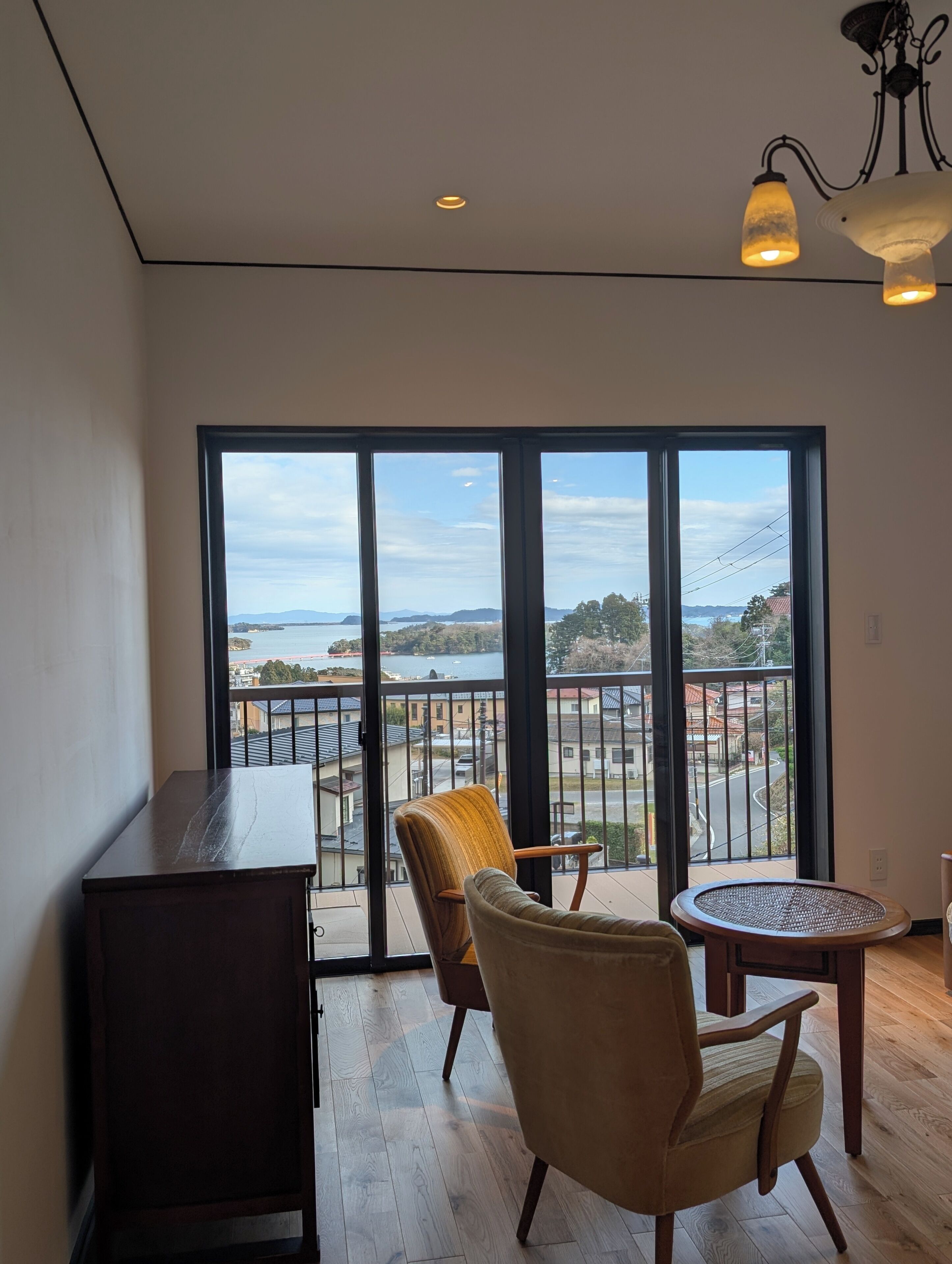 A balcony with a view of the sea, a wooden table, two chairs, and a dark wooden cabinet.
