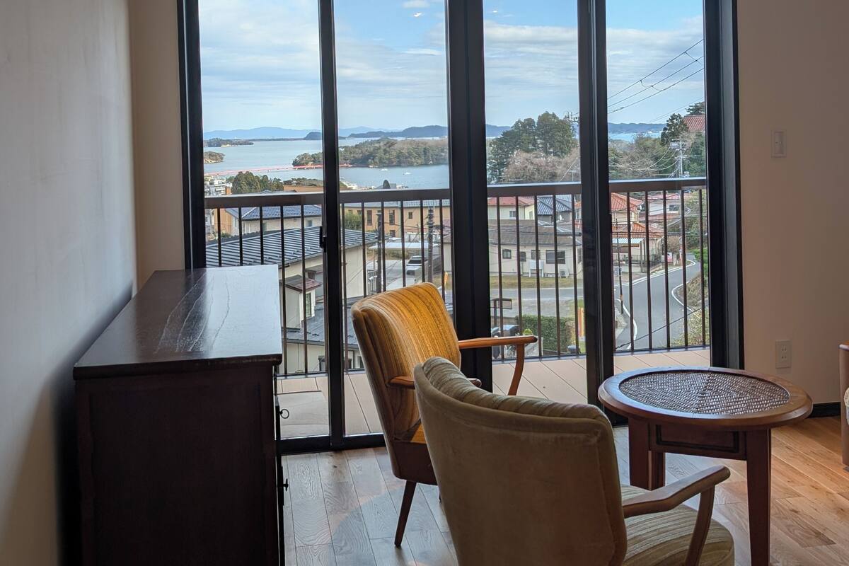 Un balcon avec vue sur la mer, une table en bois, deux chaises et un buffet en bois foncé.