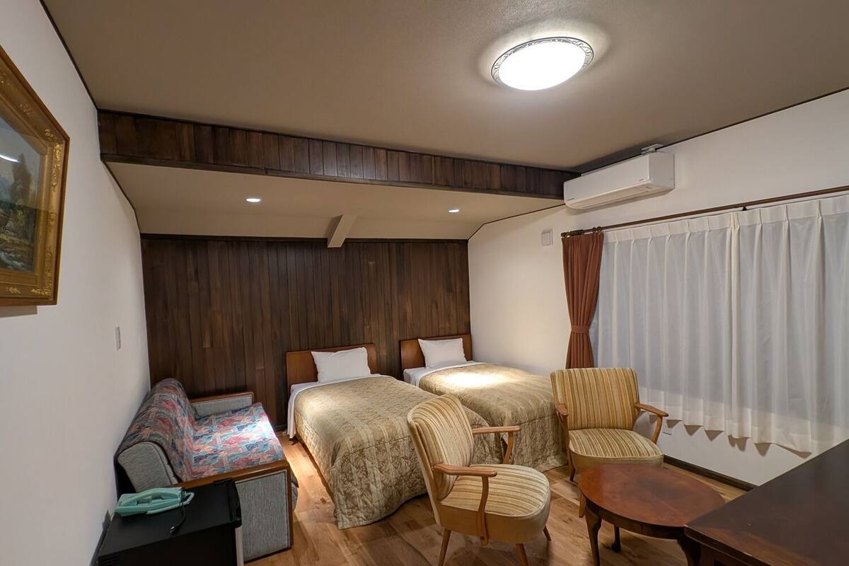 A compact hotel room with two beds, a sofa, a small table, and a chair.