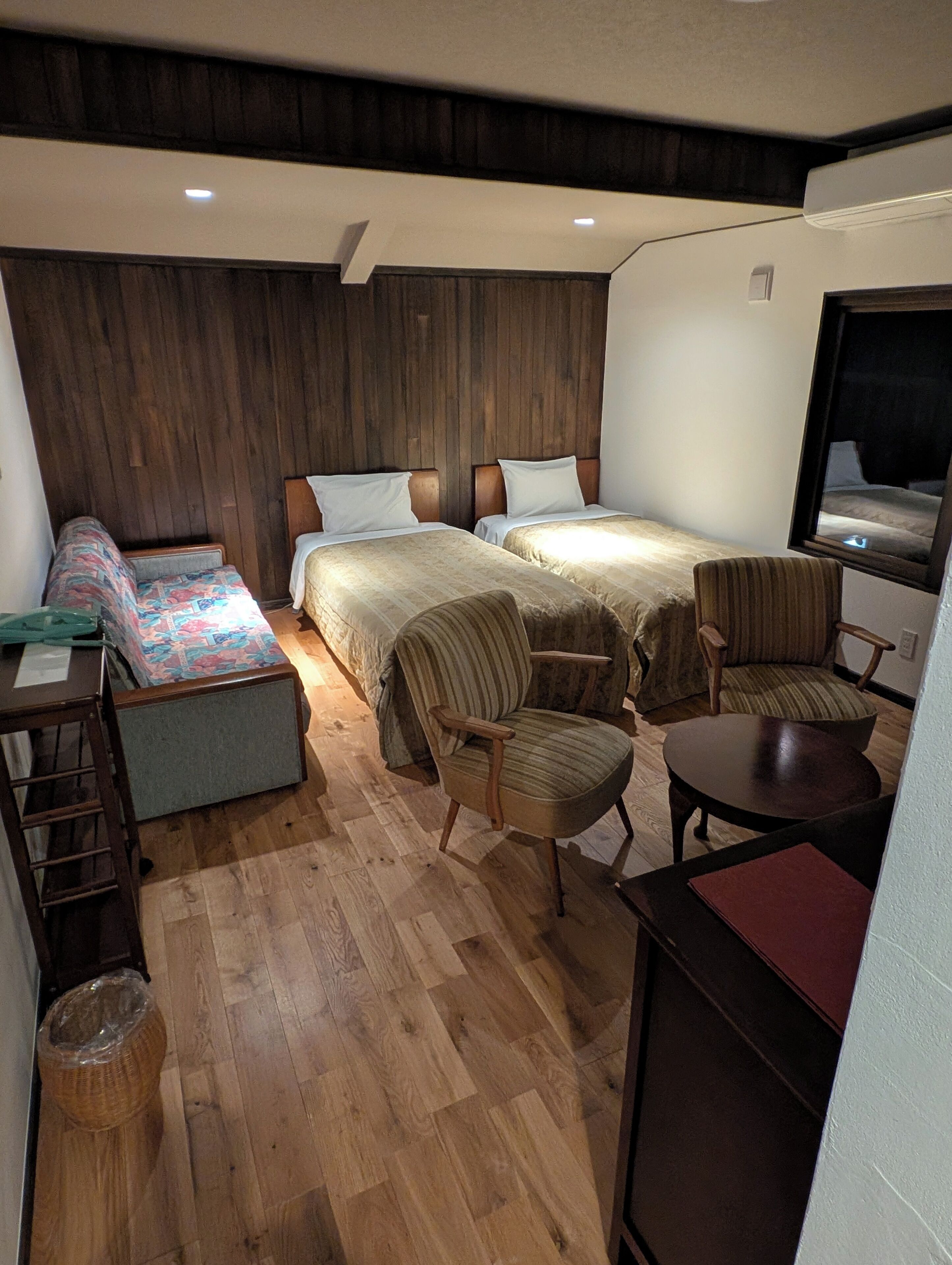 A compact hotel room with two beds, a small seating area, and a wooden floor.