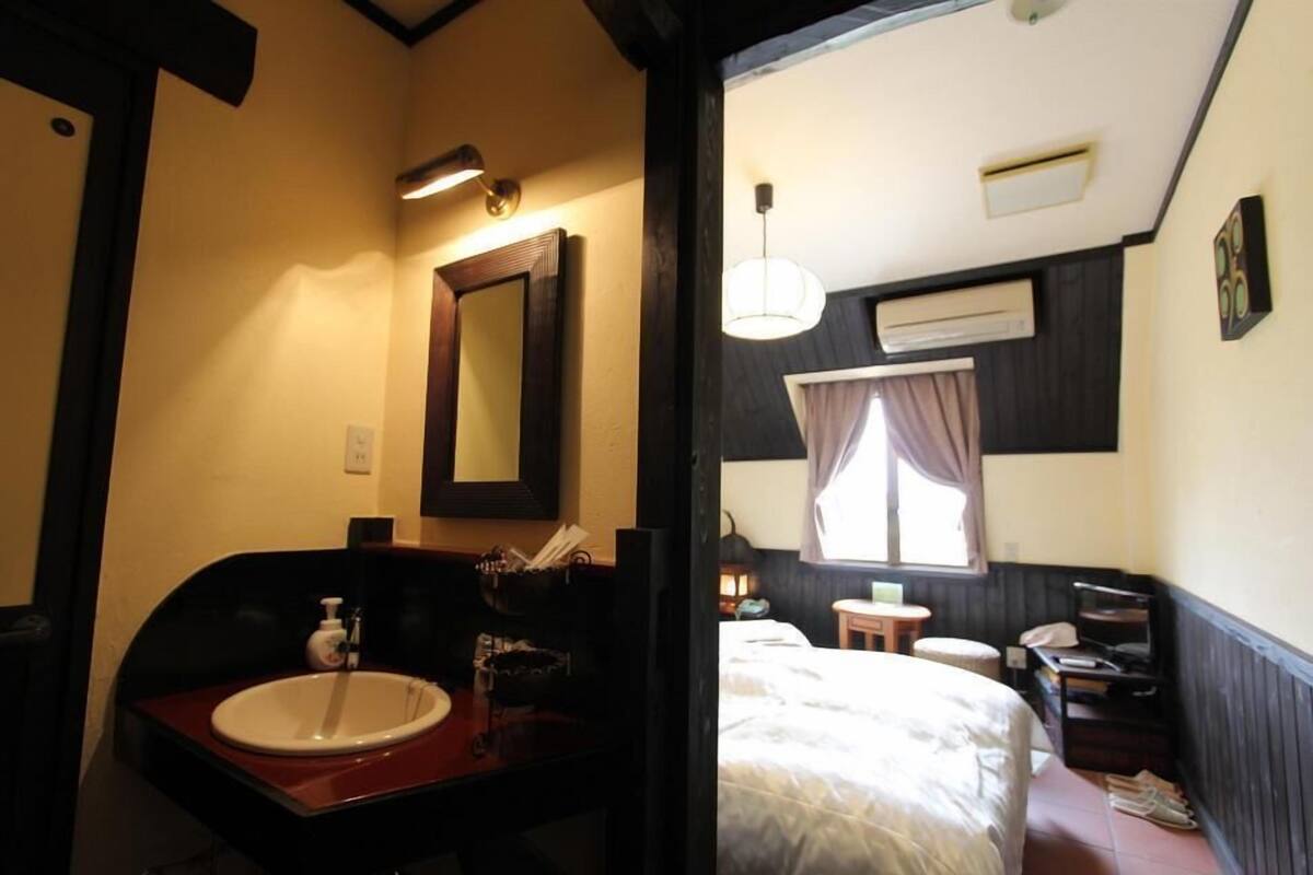 Une chambre d’hôtel comprenant un lit, une table de chevet, un petit tabouret, une fenêtre avec des rideaux, un climatiseur fixé au mur et un miroir.