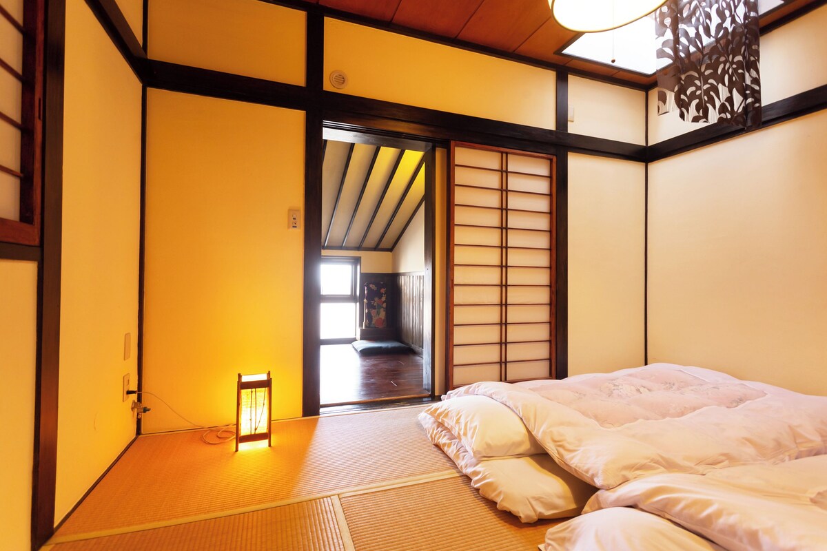 Una habitación japonesa tradicional con suelo de tatami, futón, puertas correderas shoji y vista por una puerta abierta.