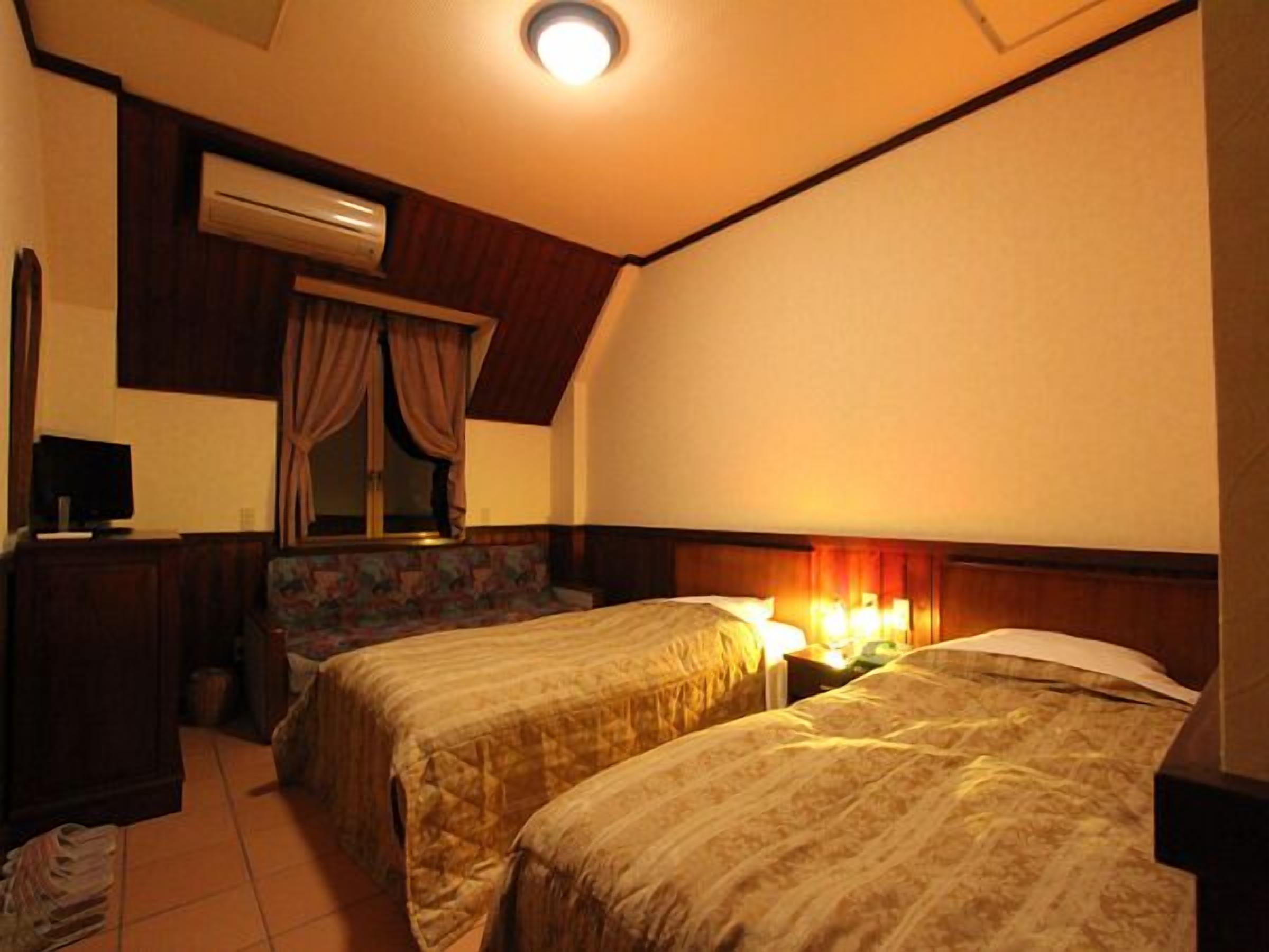 Habitació d'hotel amb dos llits, un escriptori amb un ordinador, un aparell d'aire condicionat i una finestra amb cortines.