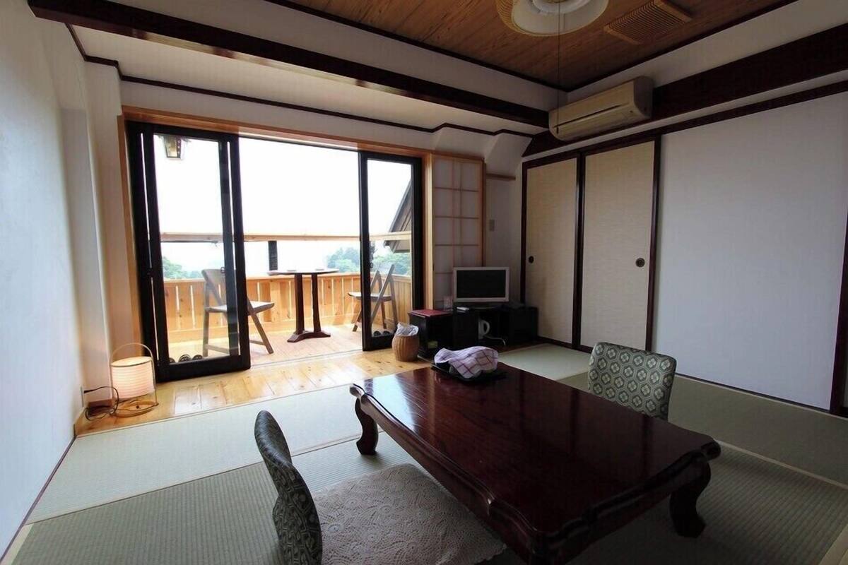 A room with tatami flooring, a wooden table, a television, sliding doors, and a view of a wooden deck outside.