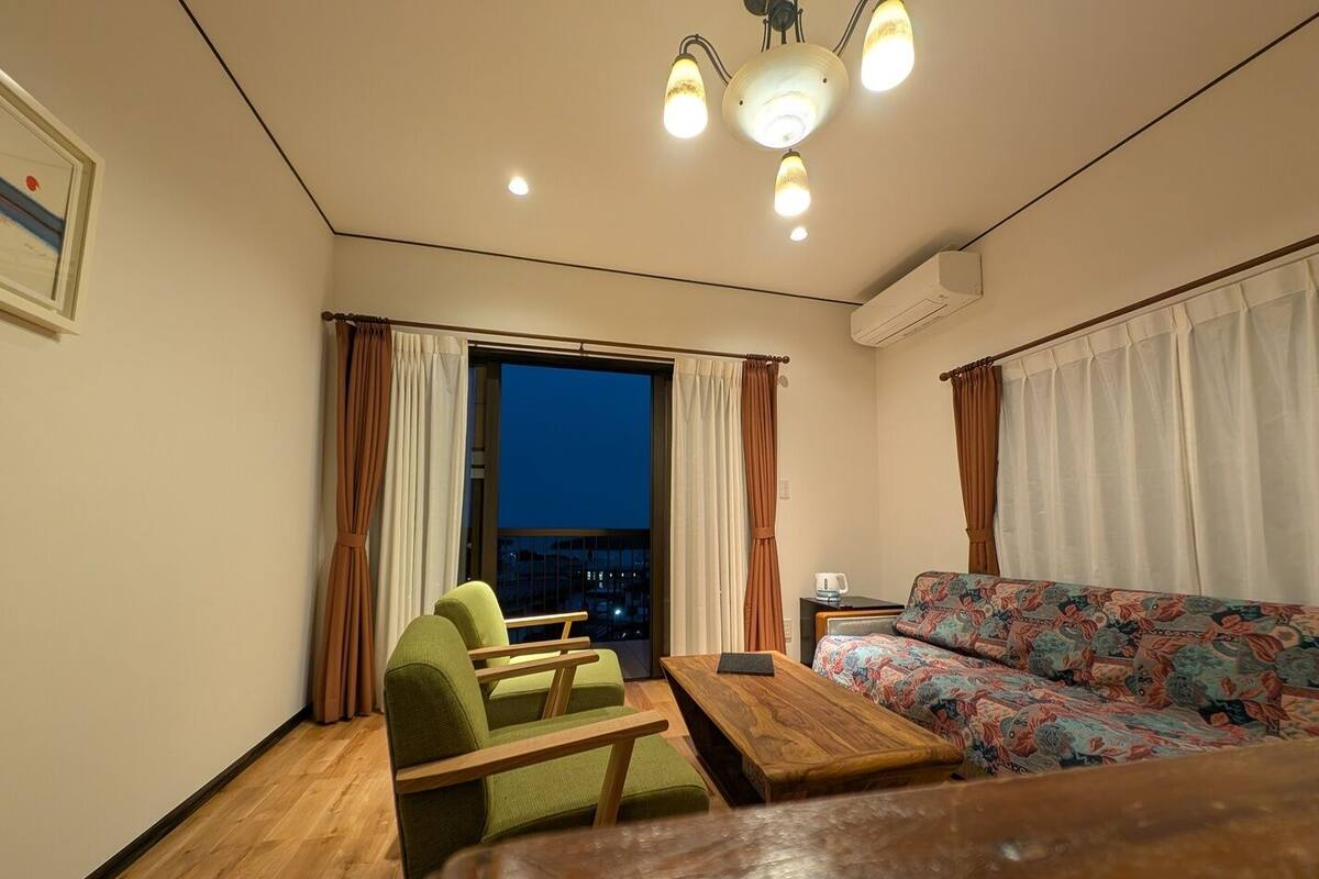Une pièce de taille modeste, comprenant un canapé, un fauteuil, une petite table et une fenêtre avec des rideaux.