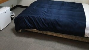 Ein Einzelbett mit dunkelblauer Bettdecke, ein kleiner weißer Kühlschrank und ein Fernseher auf einem Holzregal.