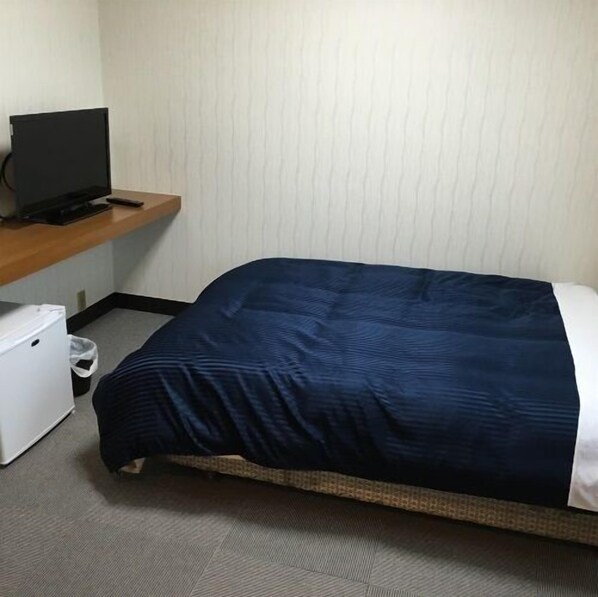A single bed with a dark blue bedspread, a small white refrigerator, and a television on a wooden shelf. - Riverine (Satsumasendai)