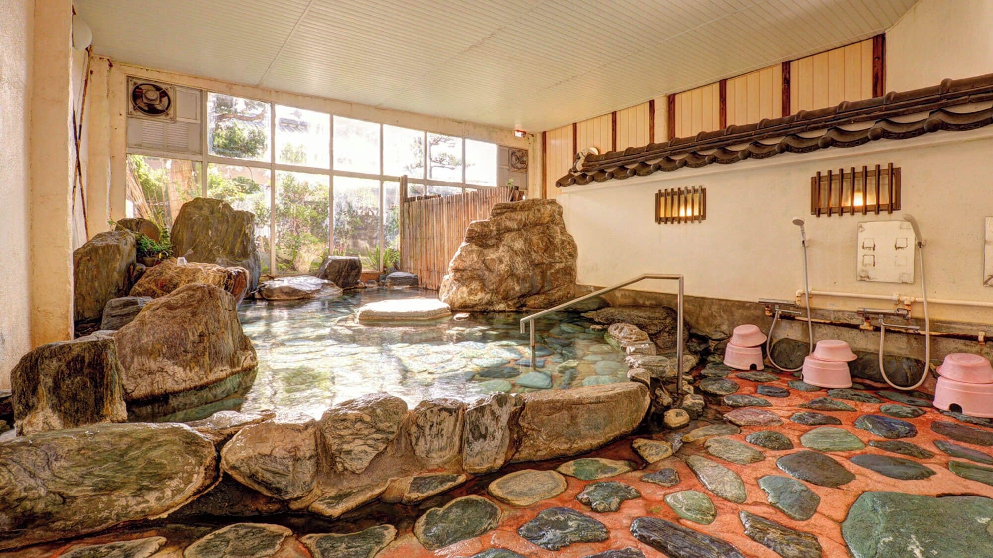 Een traditioneel Japans onsen met een stenen bad, omringd door rotsen, een houten plafond en uitzicht op groen.