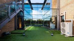 Un edificio moderno con un atrio de vidrio, un césped artificial verde y unidades de aire acondicionado exteriores.