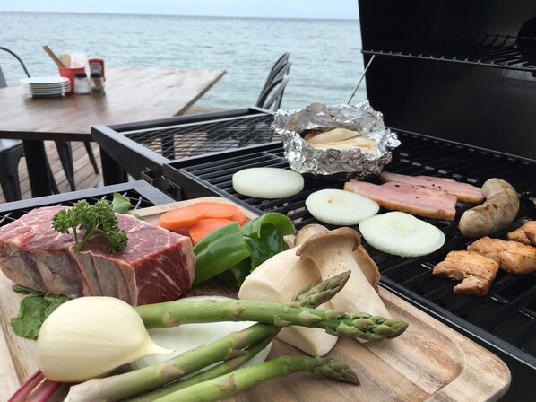 A grill with various meats and vegetables, a wooden cutting board with more vegetables, and a table with plates and condiments.