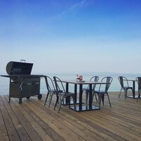 Un espacio de comedor al aire libre con una parrilla, sillas y una mesa sobre una terraza de madera con vistas al mar.