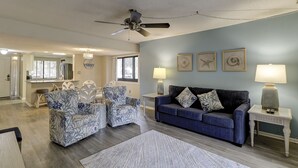 Sala de estar com um sofá azul, poltronas estampadas, um ventilador de teto e um espaço de refeições com uma cozinha ao fundo.