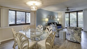 Uma sala de jantar com mesa de vidro e cadeiras brancas, um sofá com almofadas estampadas e um ventilador de teto.