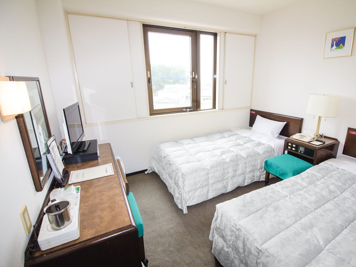 A hotel room with two beds, a desk with a computer, a window, and a nightstand with a lamp.