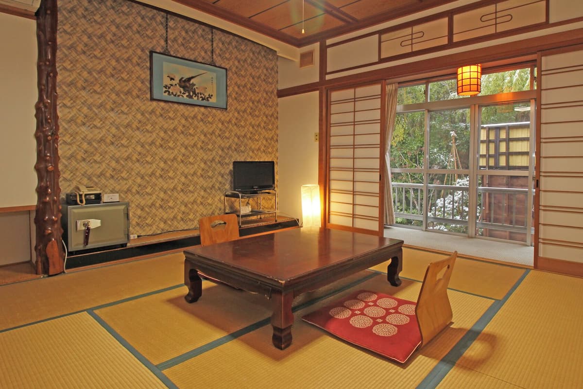 A traditional Japanese room with tatami flooring, a low table, chairs, a television, and a sliding door with a view of greenery.