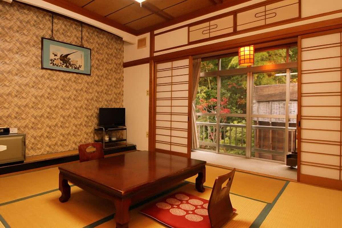 A traditional Japanese room with tatami flooring, a low table, a television, and a window with a view of greenery.