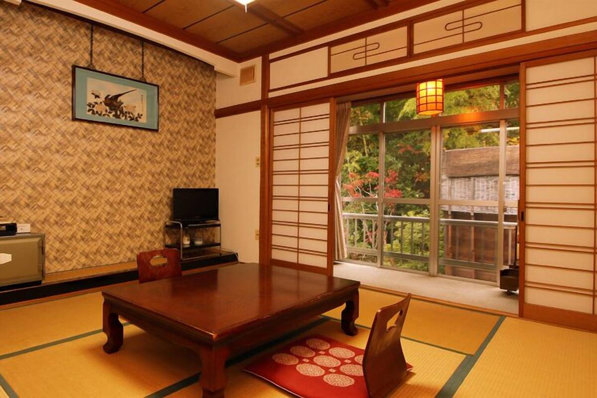 A traditional Japanese room with tatami flooring, a low table, a television, and a window with a view of greenery.