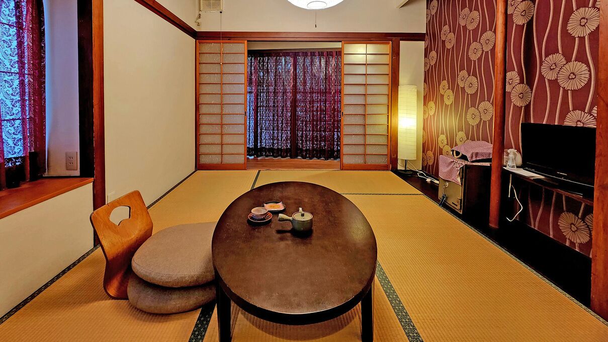Une pièce de style japonais traditionnel, avec un sol recouvert de tatamis, une petite table sur laquelle est disposé un service à thé, un fauteuil rembourré, des portes coulissantes en papier de riz (shoji) et un téléviseur posé sur un meuble en bois.