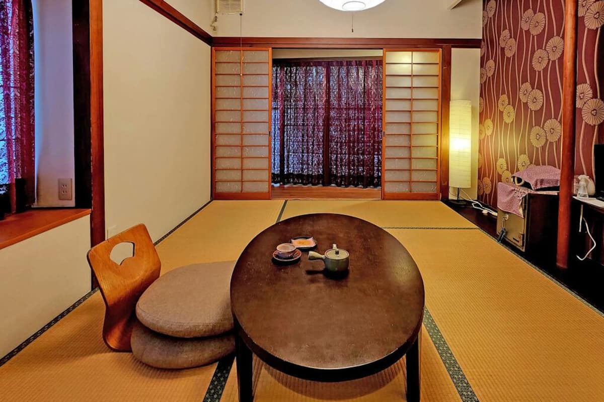 A traditional Japanese room with tatami flooring, a low table with a tea set, a cushioned chair, sliding shoji doors, and a TV on a wooden stand.