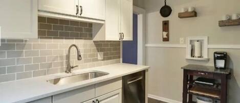 A kitchen with white cabinets, a stainless steel sink, and a dishwasher. There is a coffee maker on a shelf and a hanging pot on the wall.