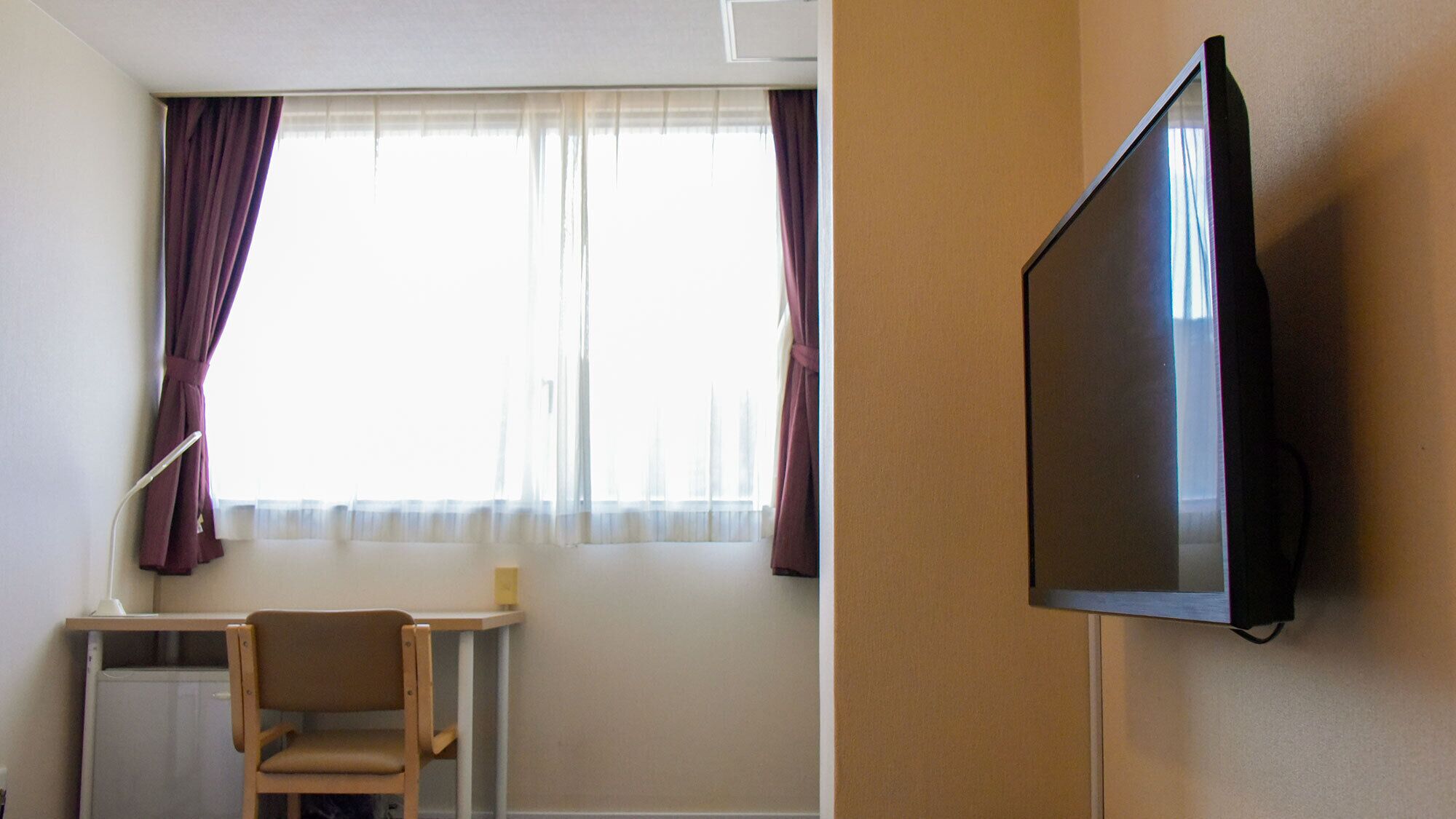 A hotel room with a bed, a TV mounted on the wall, a desk with a chair, and a window with curtains.