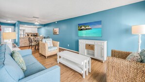 A living room with a blue wall, wooden floor, a white sofa, a white bench, wicker chairs, a fireplace, and a kitchen in the background.