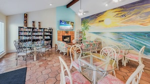 A living room with a fireplace, a large bookshelf, a dining area with white chairs, and a mural of a sunset over the ocean. - Beachside Condo | Pet-Friendly, Balcony Views & Coastal Charm (Corpus Christi)